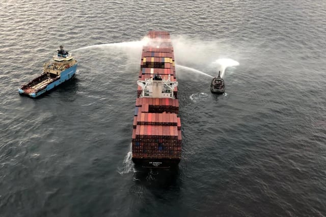 Two ships firing water cannons at another ship that has a lot of smoke pouring out of it. The smokey ship is a container ship and has lots of containers on it's deck.